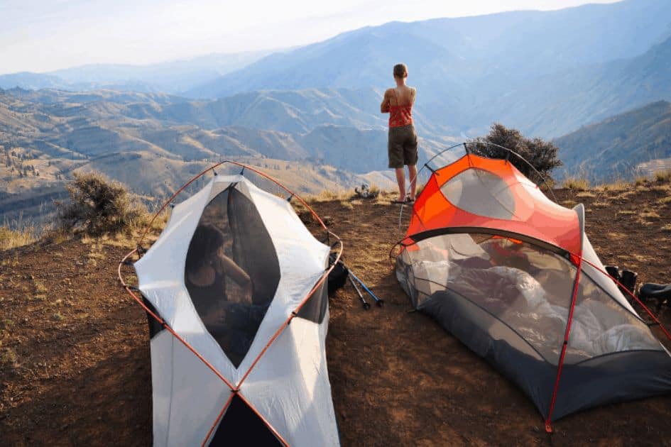 Two lightweight backpacking tents perched on a mountain top with flat ground, next to which stands a hiker admiring the panoramic view.